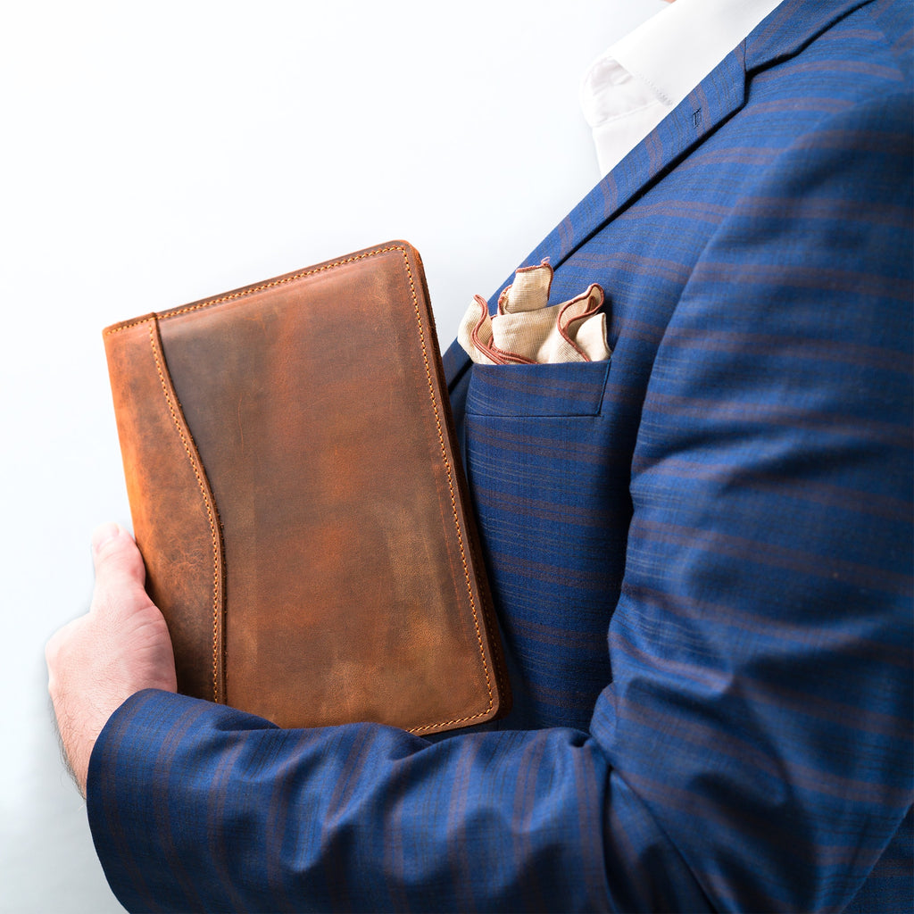 Man in a navy pinstripe suit holding a closed PEGAI Eriksen Jr. padfolio in mahogany Dallas Crazy Horse leather under his arm, pocket square visible.