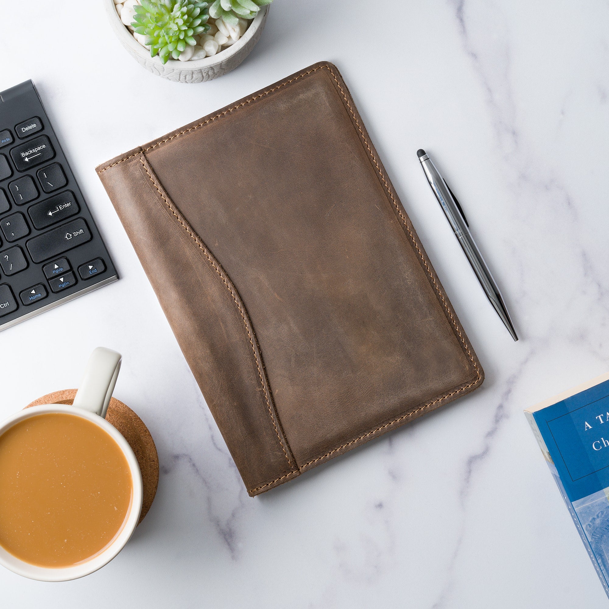 Sand Brown Eriksen padfolio lies flat on a marble desk beside a keyboard, succulent, pen, coffee cup, and book, styled for an office setting.