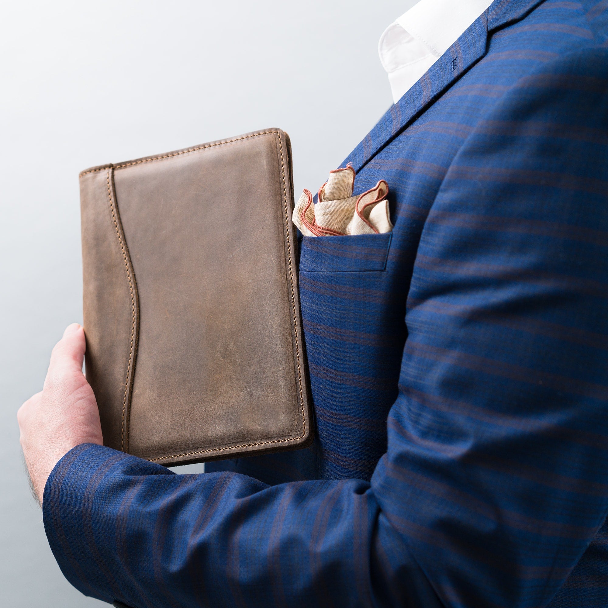 Man in a blue pinstripe suit holds the closed Sand Brown Eriksen padfolio against his chest, pocket square visible in his jacket pocket.