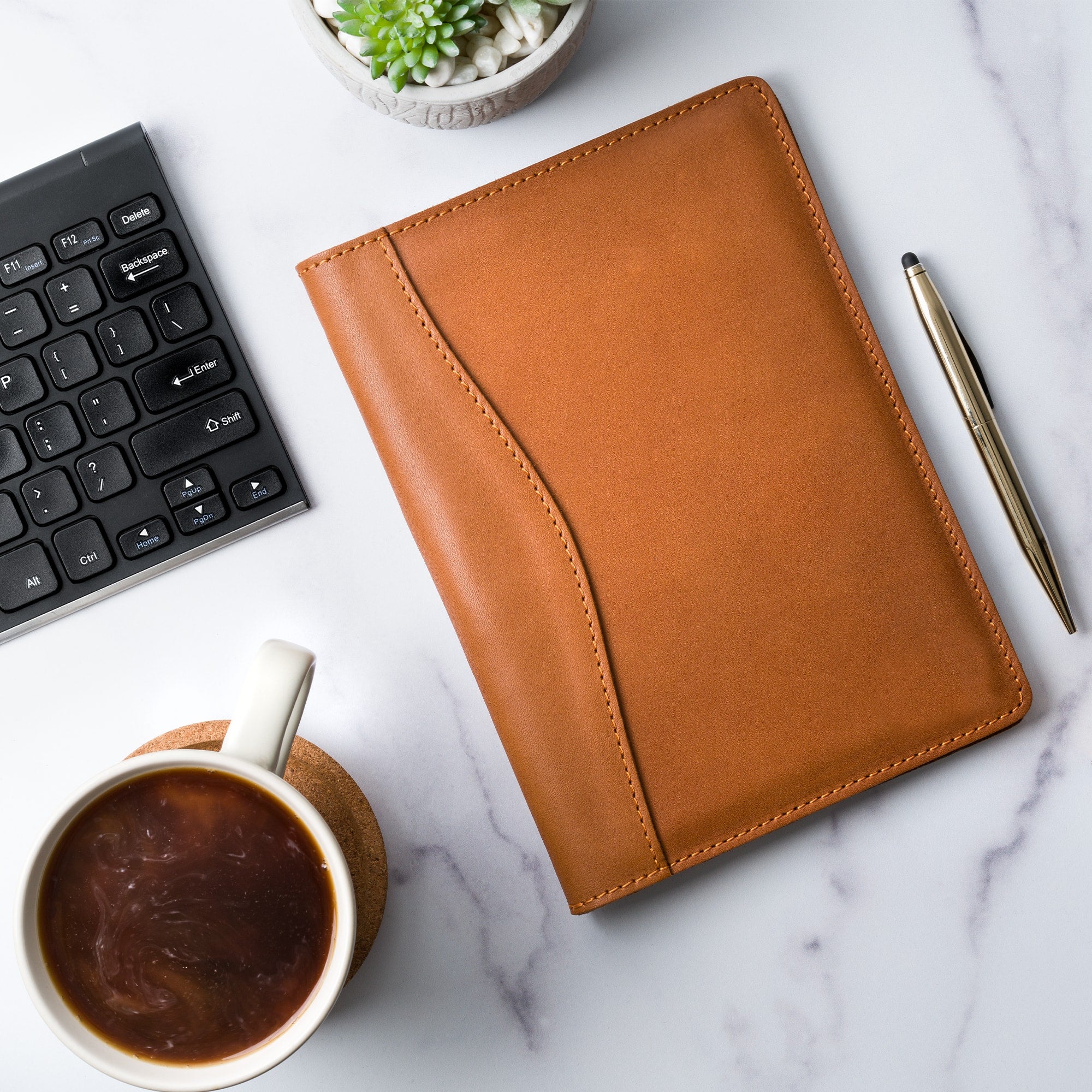 PEGAI Eriksen Jr. padfolio in cognac brown Zurich calfskin lying closed on a marble desk beside a keyboard, stylus pen, succulent, and a cup of coffee.
