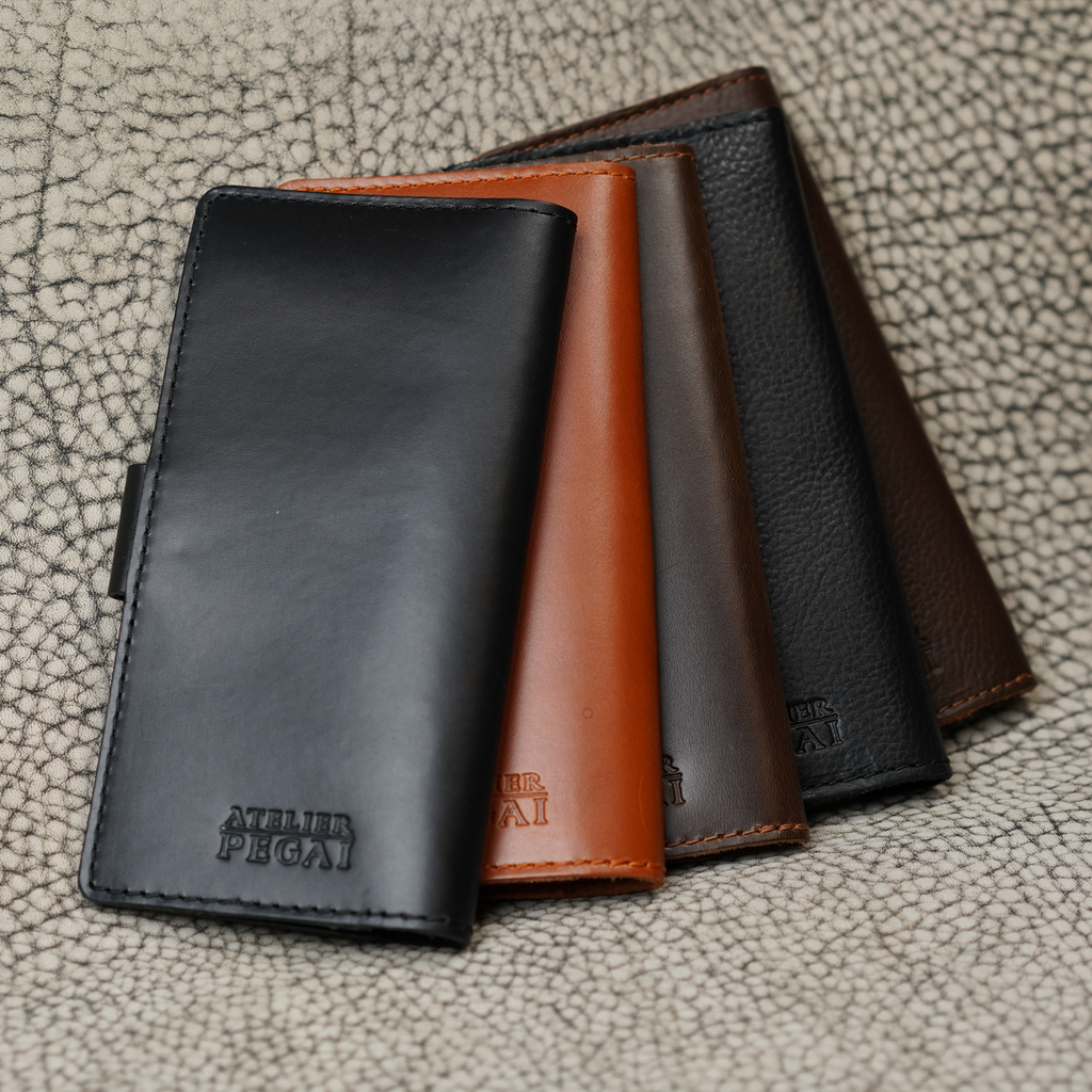 Five Clark Checkbook Covers fanned out on crackled leather: black, whiskey, coffee, Dymnos Black, and Dymnos Brown. All show the debossed Atelier PEGAI logo. Showcases the new full Clark colorway range.