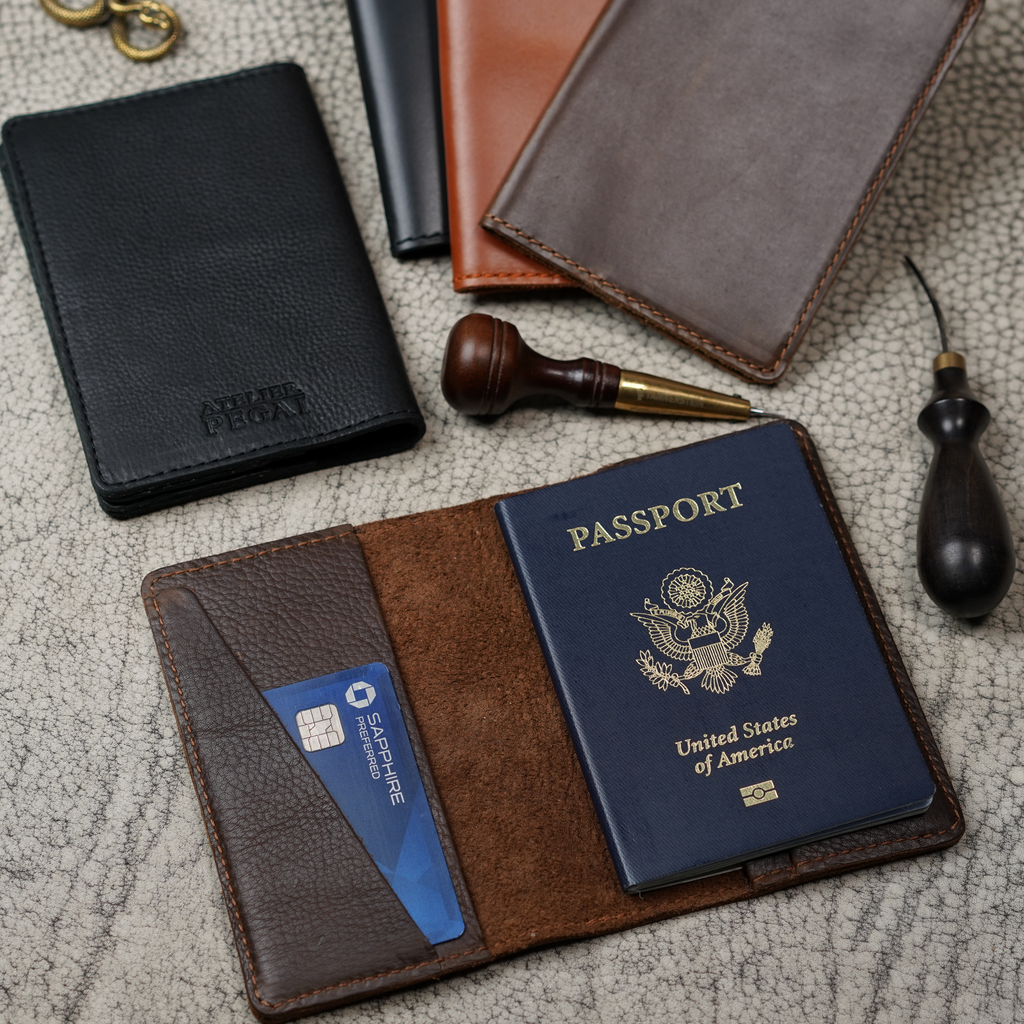 Editorial flat lay showing multiple passport covers and leather goods on a crackle-textured surface. Center: open brown leather passport cover with a U.S. passport and credit card inserted. Upper left: black Atelier PEGAI card case. Craftsman tools and tan passport cover in background.