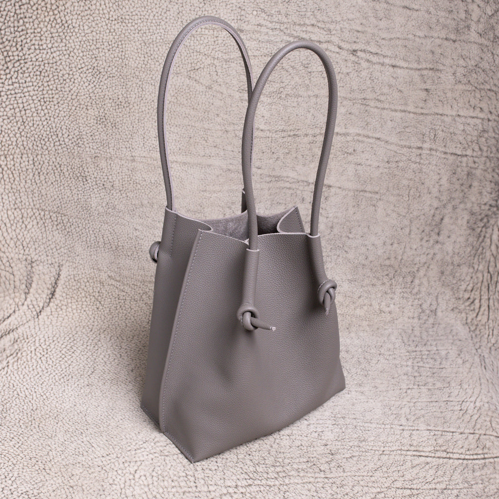 Three-quarter angle of the gray Flora tote, highlighting its sculpted silhouette and rolled handles against a neutral textured backdrop.