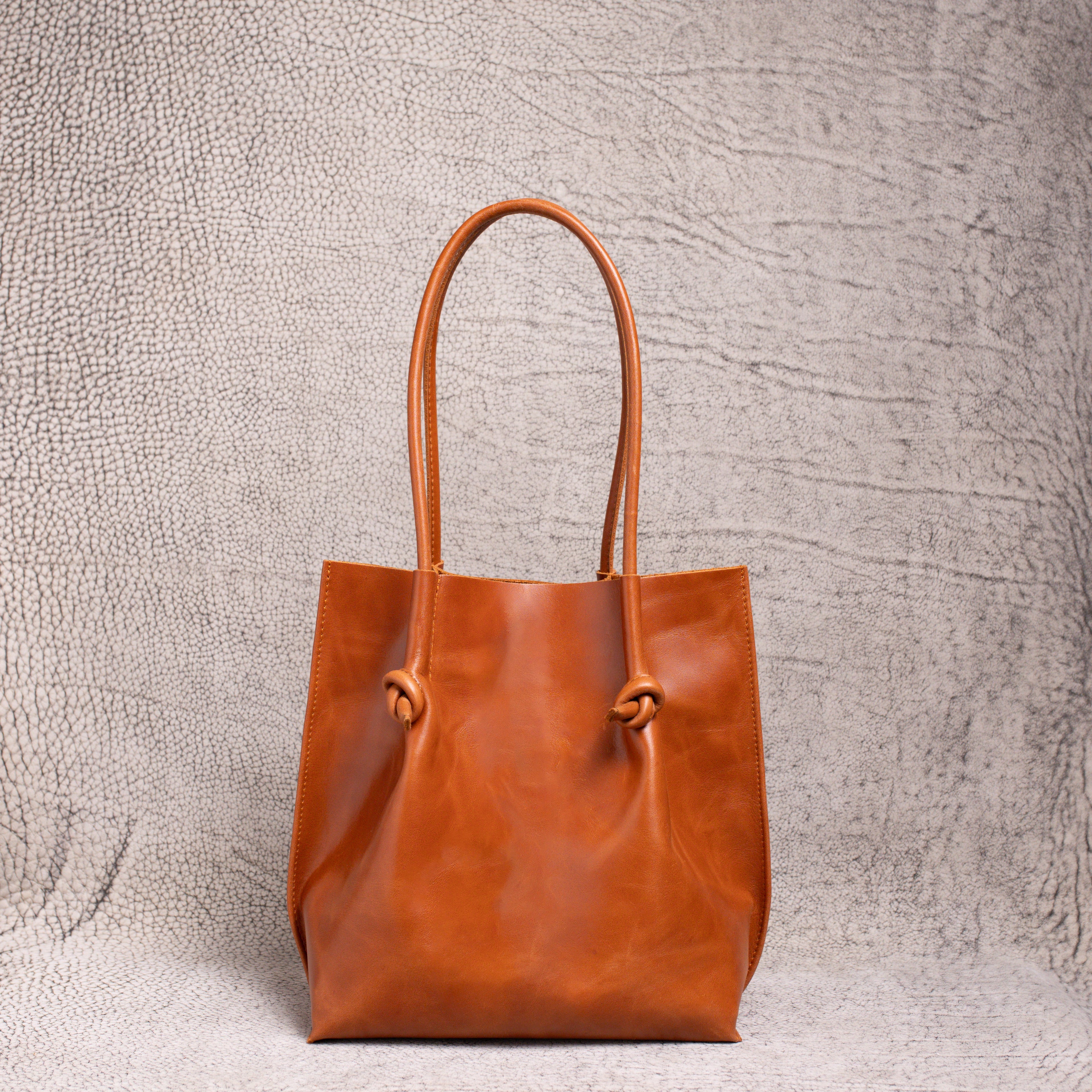A front-facing view of the Flora Everyday Leather Tote in Dual Tone Whiskey, showcasing its minimalist silhouette and knotted leather handles.
