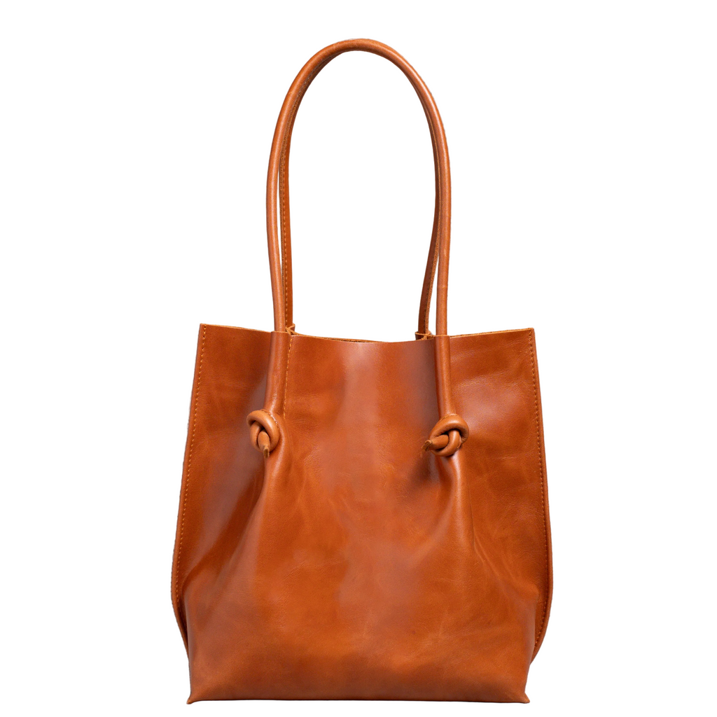 Flora Everyday Tote in Dual Tone Whiskey leather stands upright, tall silhouette, with rolled dual handles and a single knotted leather closure on each side of the open top.
