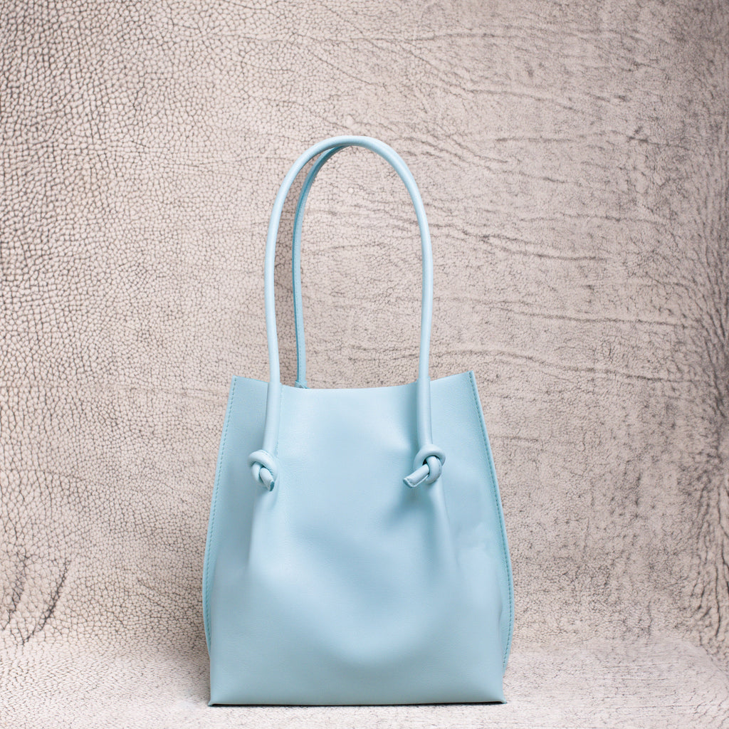 A minimalist light blue leather tote bag with knotted handle detailing, standing upright against a textured neutral backdrop.