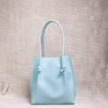 A minimalist light blue leather tote bag with knotted handle detailing, standing upright against a textured neutral backdrop.