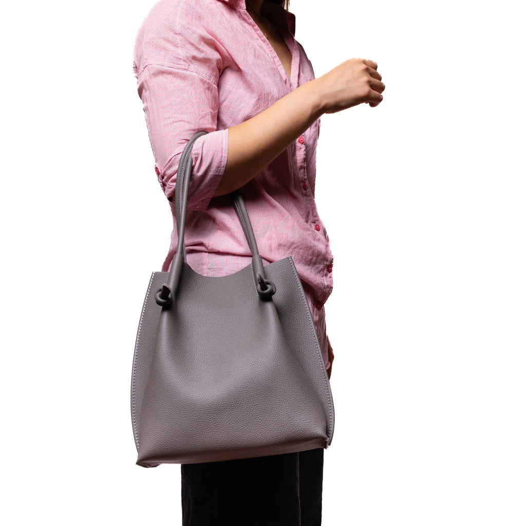 Model wearing a pink blouse carries the Flora Everyday Tote in gray, showing its size and elegant shape in a lifestyle setting.
