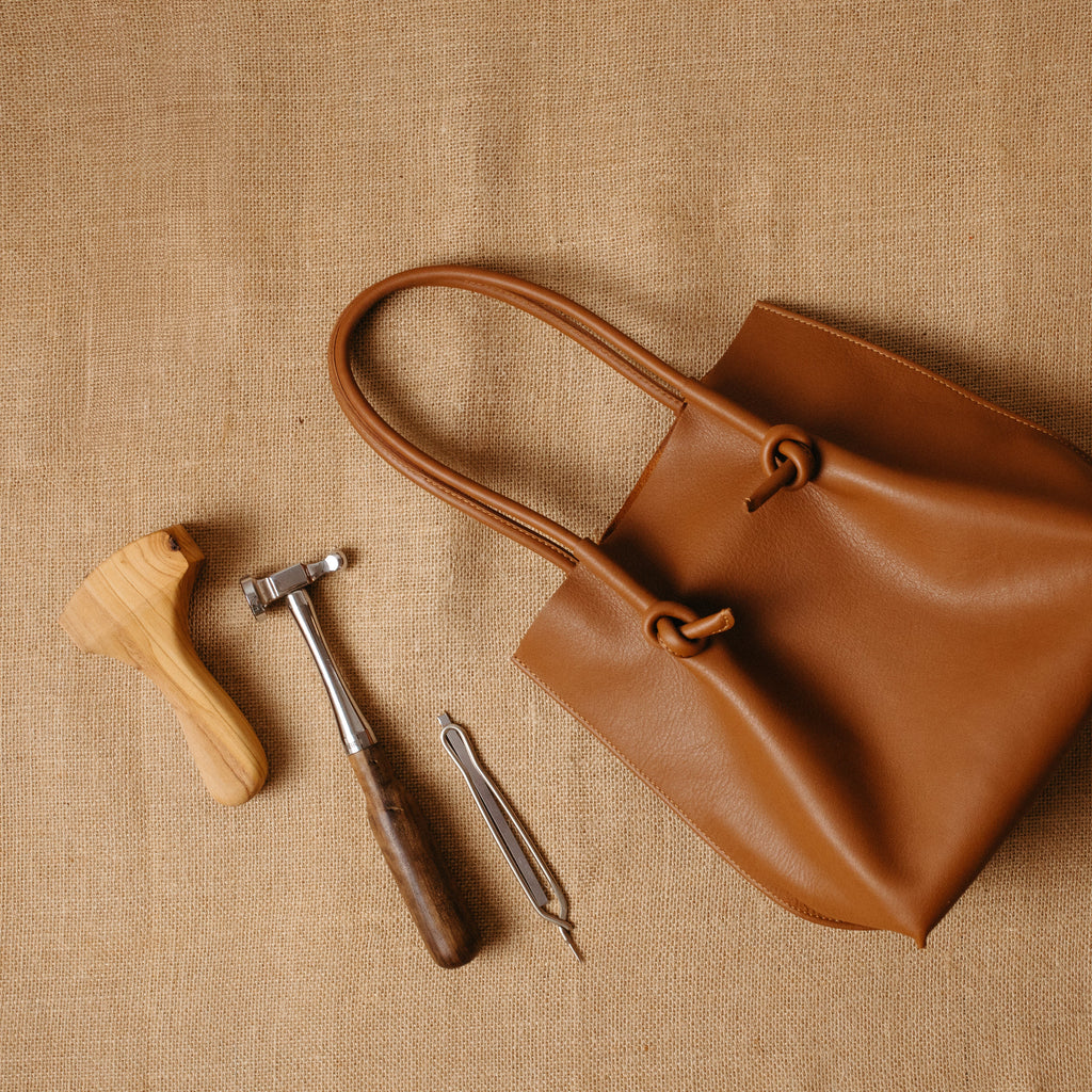 The tan (taba) Flora tall tote bag laid on a burlap surface next to traditional leathercraft tools, representing handmade quality and artisan craftsmanship.