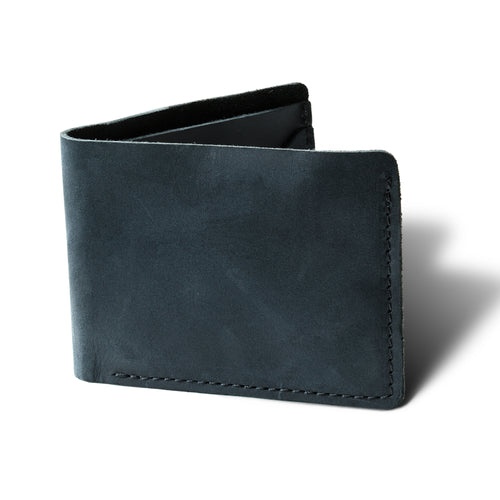 Bureau Leather Bifold Wallet in Charcoal Black, standing upright at a three-quarter angle on a white surface. Dallas Crazy Horse calfskin in a deep slate charcoal tone with natural waxy surface variation, pull-up depth, and subtle hide markings. Tonal dark saddle stitching runs along the right edge and bottom. Wallet is slightly open, revealing a dark suede interior spine.
