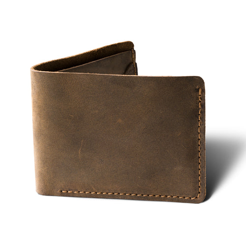 Bureau Leather Bifold Wallet in Chestnut Brown, standing upright at a three-quarter angle on a white surface, shown without personalization. Dallas Crazy Horse calfskin in a warm dusty chestnut tone with natural waxy surface variation, visible hide scratches, and pull-up depth. Warm tonal saddle stitching runs along the right edge and bottom. Raw suede spine partially visible at the top fold.