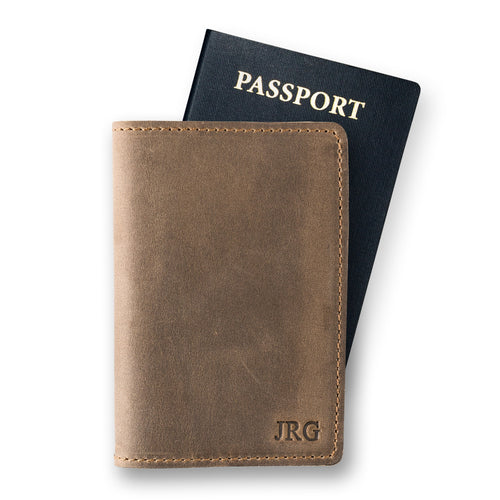 Sand brown leather passport cover in front of a navy U.S. passport on a white background. Waxy crazy horse surface with warm taupe tonal variation. Fire-branded initials "JRG" embossed in serif font at the lower front panel. Tan hand-stitched perimeter visible along right edge.