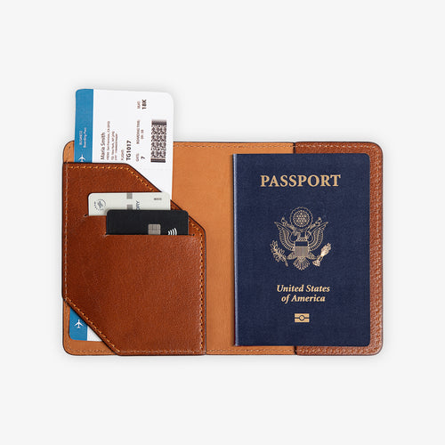 Full interior view of the open Phokion brown leather passport wallet, fully loaded. Left panel shows the diagonal-cut card pocket with two cards and a boarding pass fanned out above. Right panel holds a full U.S. passport. Warm cognac tan leather lining with tonal brown saddle stitching.