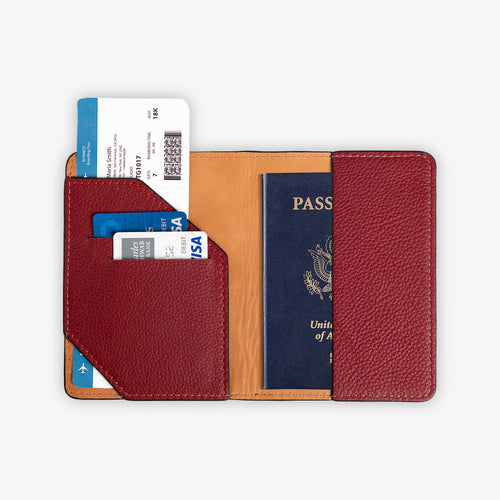 Full interior view of the open burgundy leather passport wallet, fully loaded. Left panel shows the diagonal-cut burgundy card pocket with two debit cards and a boarding pass fanned out above. Right panel holds a U.S. passport. Warm cognac tan leather lining and red stitching visible.