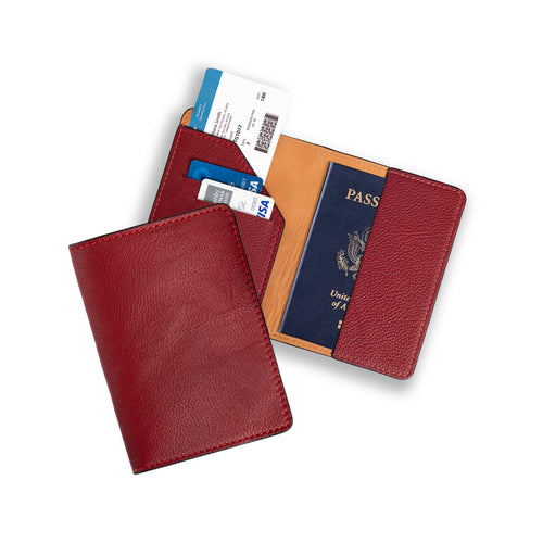 Two burgundy leather passport wallets on white: one closed showing the pebbled exterior, one open displaying the warm tan lining, diagonal card pocket with two debit cards and a boarding pass on the left, and a U.S. passport inserted on the right. Red contrast stitching throughout.