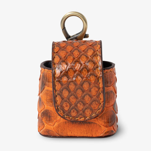 Coil AirPods case in Sunset python leather, angled side view. Vibrant orange and deep brown scales with golden contrast stitching detail. Curved magnetic closure with dark trim. Antique brass clip hardware.