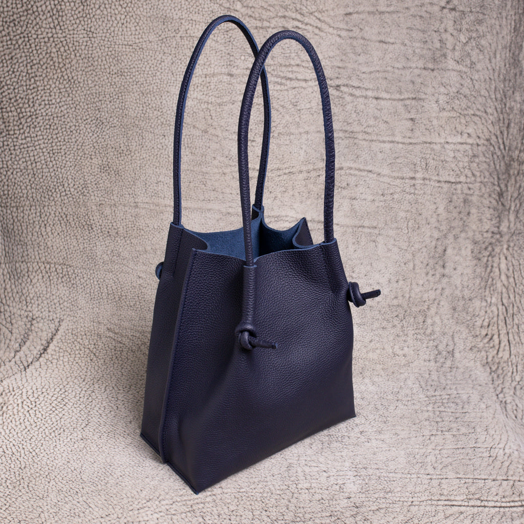Handcrafted Flora Leather Tote in Togo Blue—shown in an upright three-quarter view. Soft-grained Togo leather, knotted rolled handles, and raw suede interior subtly visible.