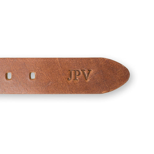 Close-up of the tip end of the Buffalo Leather Belt in Saddle Brown showing fire-branded personalization. Three bold initials "JPV" embossed in Times New Roman near the belt tip, set against warm saddle-toned buffalo hide with visible natural grain variation and adjustment holes along the edge.