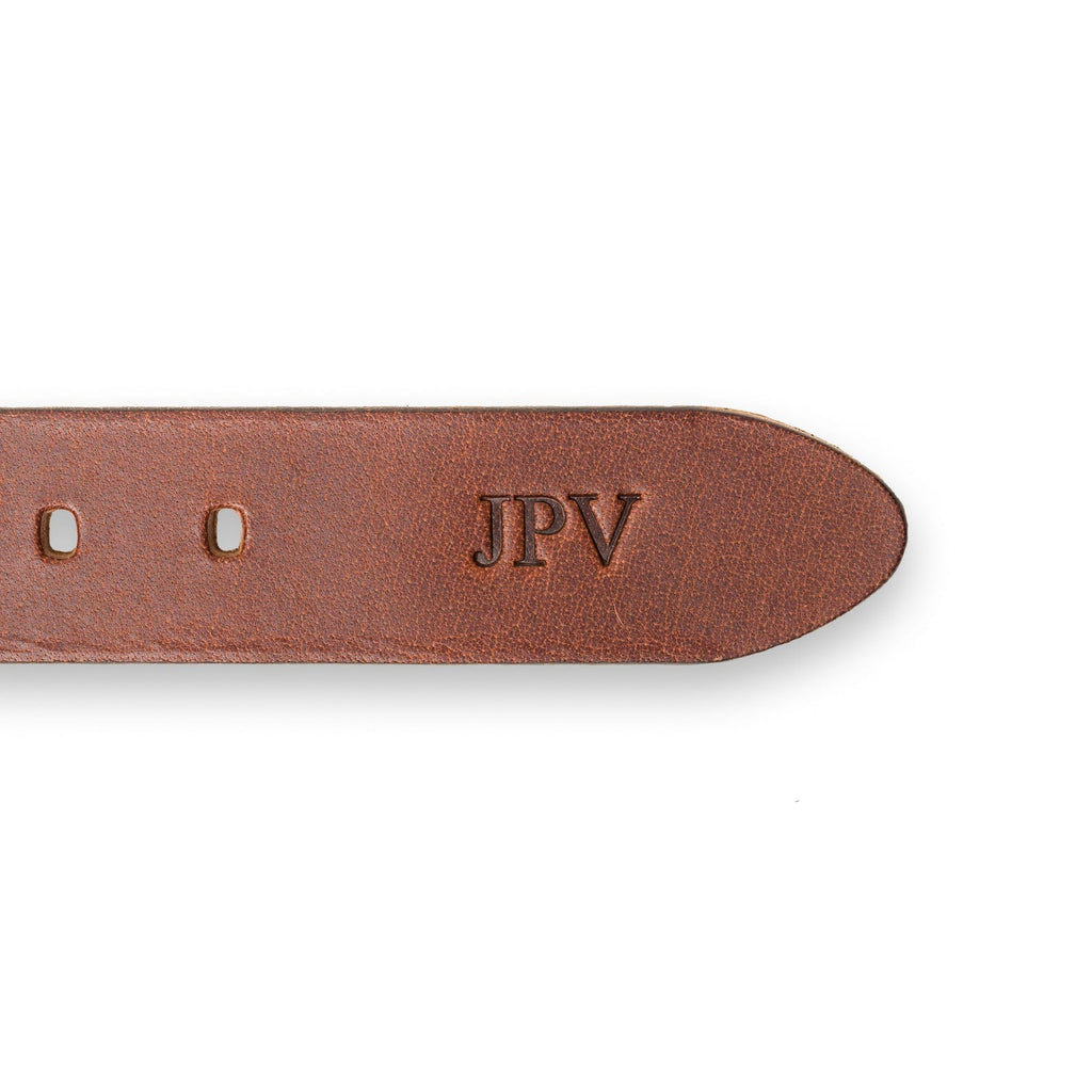 Close-up of the tip end of the Buffalo Leather Belt in Rustic Brown showing fire-branded personalization. Three bold initials "JPV" embossed in Times New Roman near the belt tip, set against warm russet calfskin with visible natural grain texture and adjustment holes along the edge.