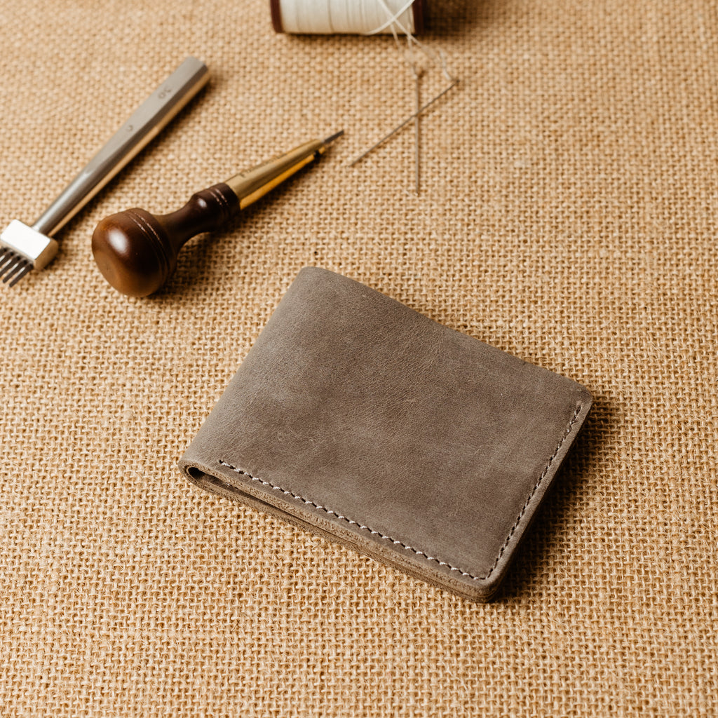 Bureau Leather Bifold Wallet in Rock Gray resting closed on a natural burlap surface, showing the clean exterior back face. Cool slate gray Dallas Crazy Horse calfskin with characteristic waxy pull-up surface, natural hide markings, and subtle tonal depth. Artisan leather tools — stitching chisel, wooden-handled awl, thread spool, and needles — arranged in the background.