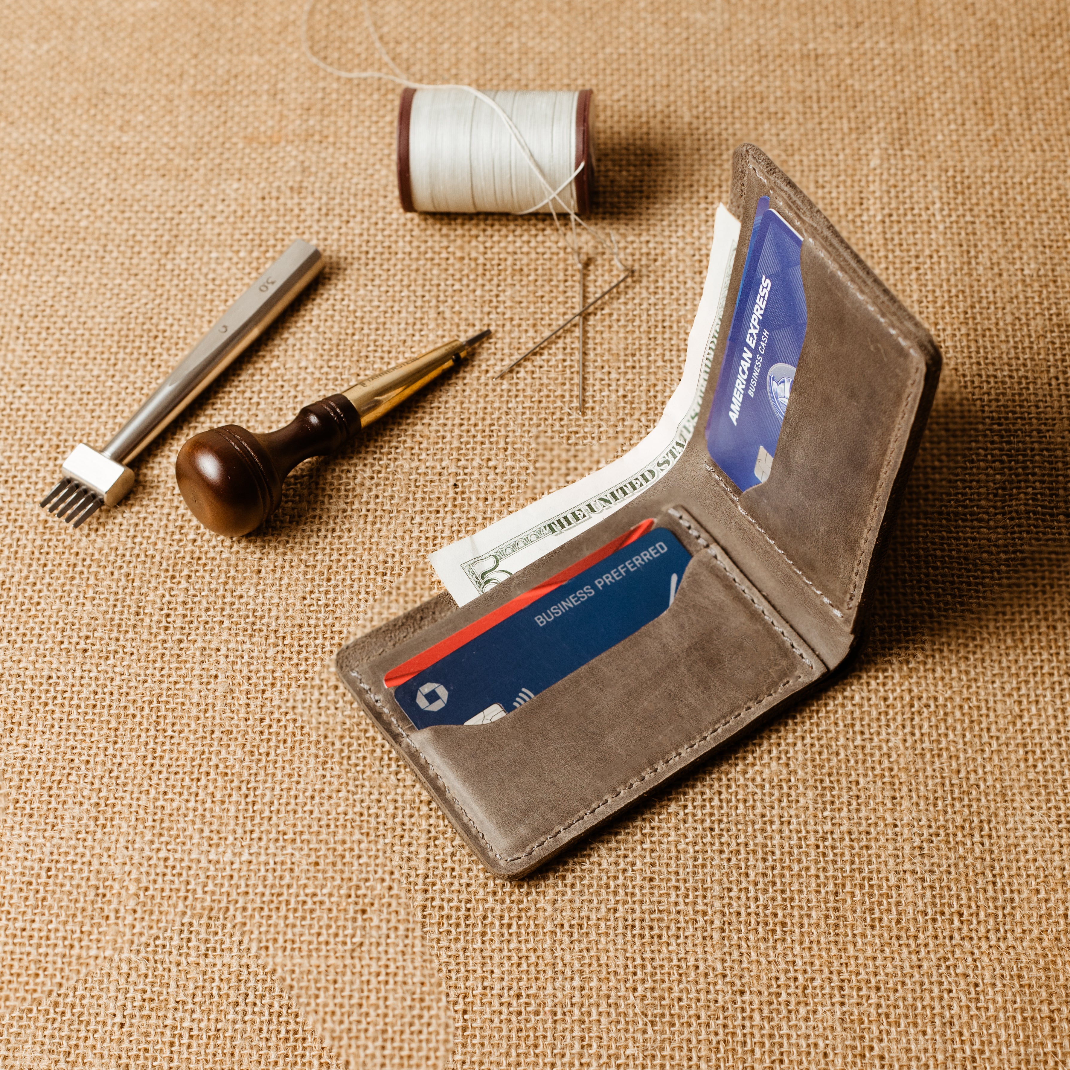Bureau Leather Bifold Wallet in Rock Gray opened at an angle on a natural burlap surface, loaded with an American Express card, a Chase Ink Business Preferred card, and a folded US dollar bill. Four card slots and one bill compartment clearly visible. Cool gray Dallas Crazy Horse calfskin interior with natural tonal variation. Artisan leather tools — stitching chisel, wooden-handled awl, thread spool, and needles — arranged in the upper left background.