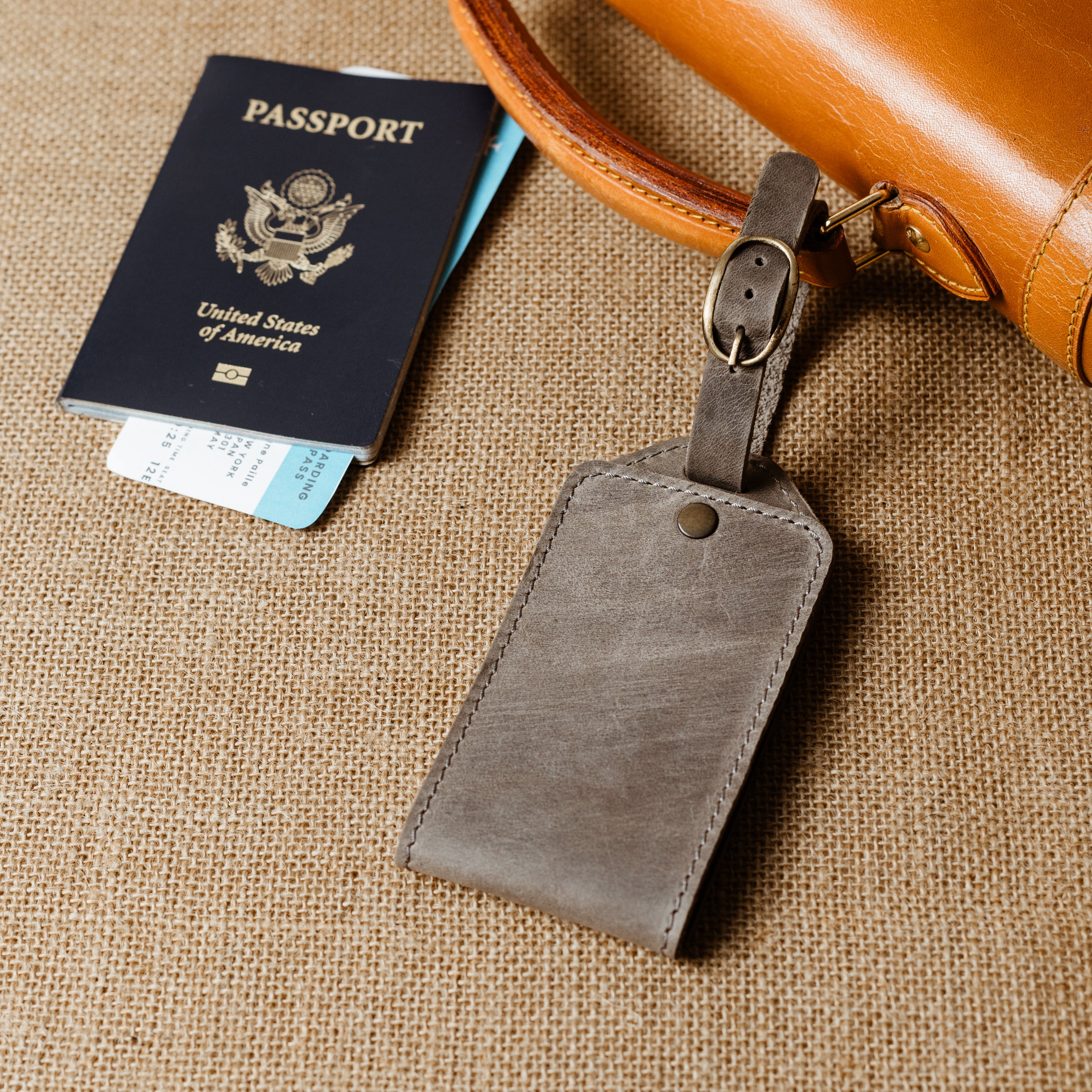 Personalized gray leather luggage tag attached to carry-on bag beside a passport