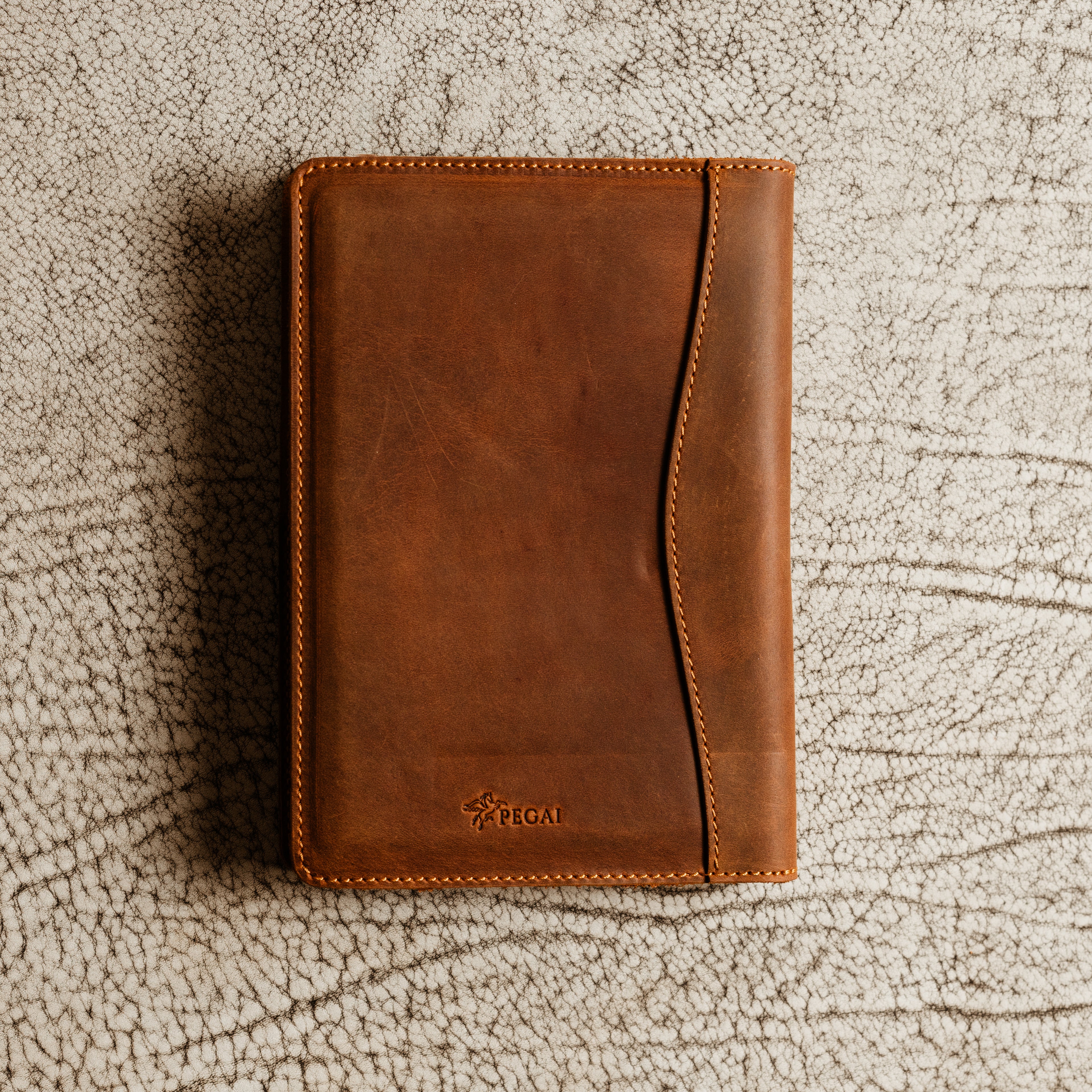 PEGAI Eriksen Jr. padfolio in mahogany Dallas Crazy Horse leather lying closed on a textured white surface, showing PEGAI pegasus logo and curved seam stitching. 
