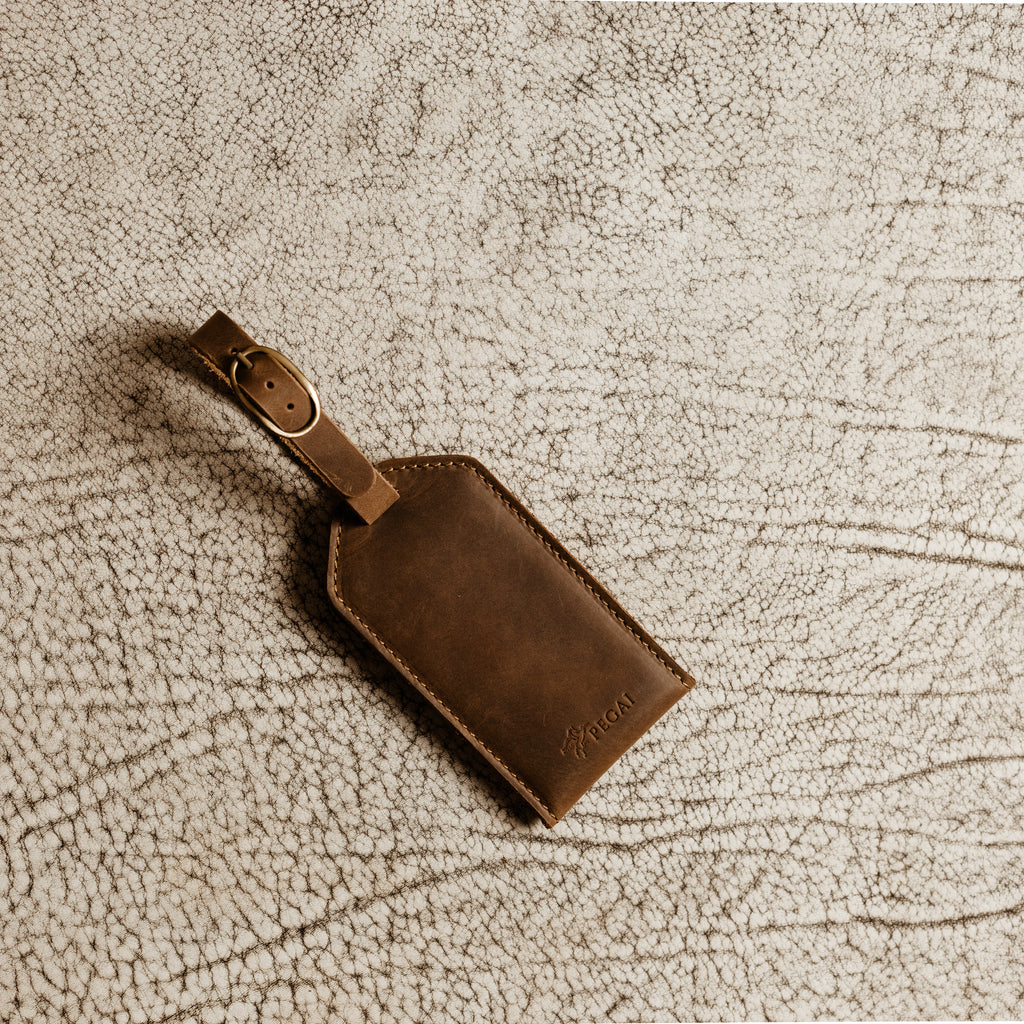 Brown leather luggage tag with antique brass buckle resting on textured leather surface