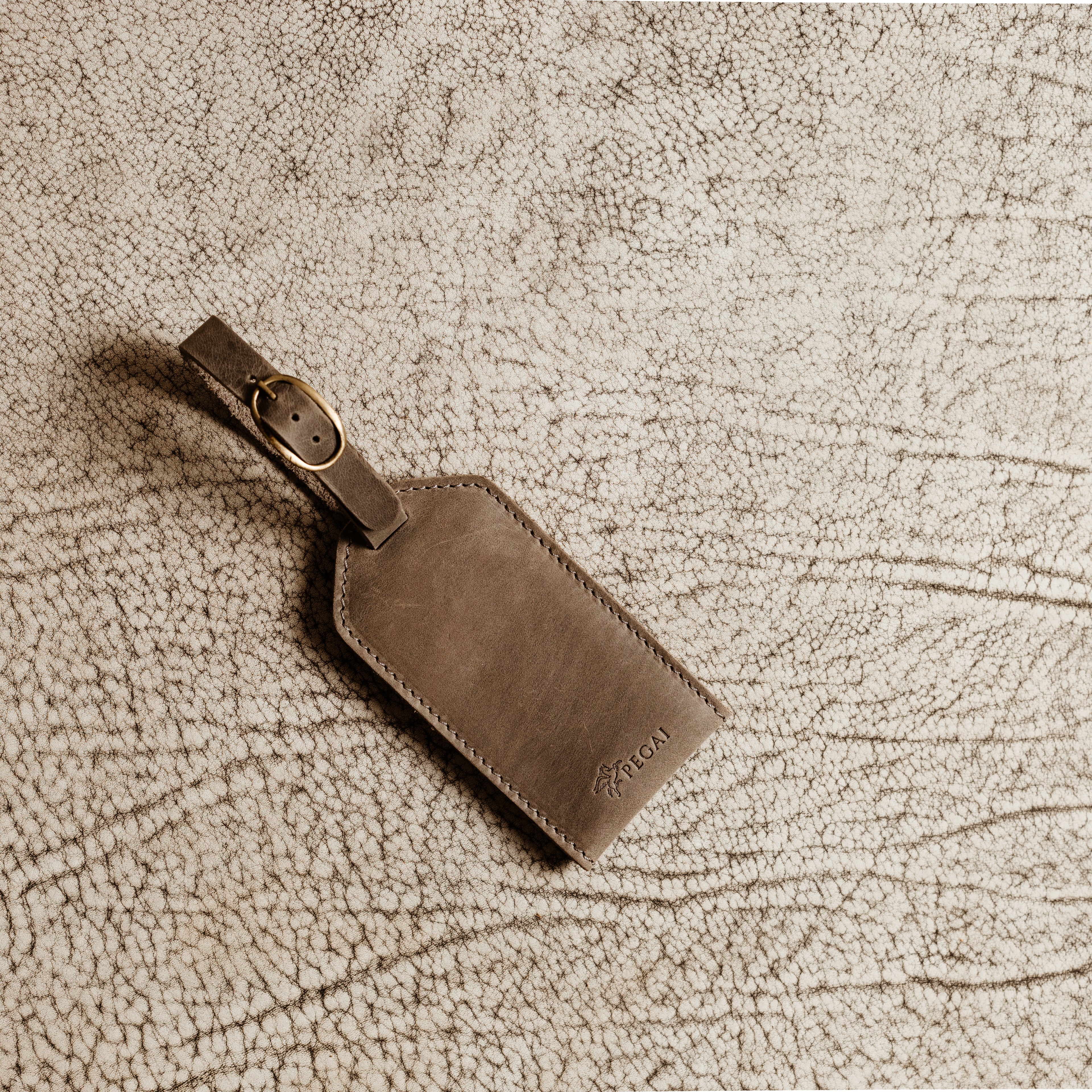 Gray leather luggage tag with antique brass buckle resting on textured fabric background