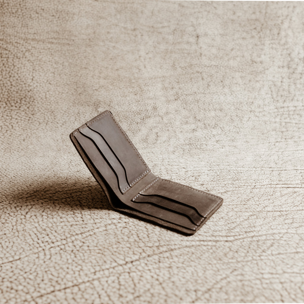 Bureau Leather Bifold Wallet in Rock Gray opened wide and standing upright on a cream crackled leather surface under warm directional lighting. Interior fully visible: four angled card slot openings across both panels, separated by a central bill compartment. Cool gray Dallas Crazy Horse calfskin interior with rich tonal variation and tonal gray saddle stitching throughout.