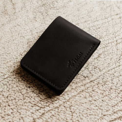 Bureau Leather Bifold Wallet in Charcoal Black resting closed on a cream-toned crackled leather surface. Deep slate charcoal Dallas Crazy Horse calfskin exterior with natural waxy sheen and subtle tonal variation. Debossed PEGAI horse logo and wordmark visible on the lower right corner of the front face. Tonal dark saddle stitching along the right edge.