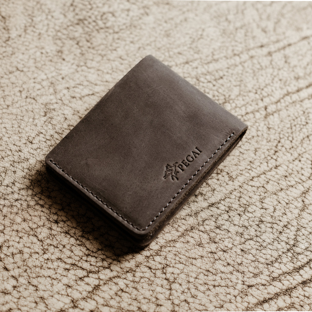 Bureau Leather Bifold Wallet in Rock Gray resting closed on a cream-toned crackled leather surface. Deep cool gray Dallas Crazy Horse calfskin exterior with natural waxy sheen and subtle tonal cloudiness. Debossed PEGAI Pegasus logo and wordmark visible on the lower right corner. Tonal gray saddle stitching along the right edge.