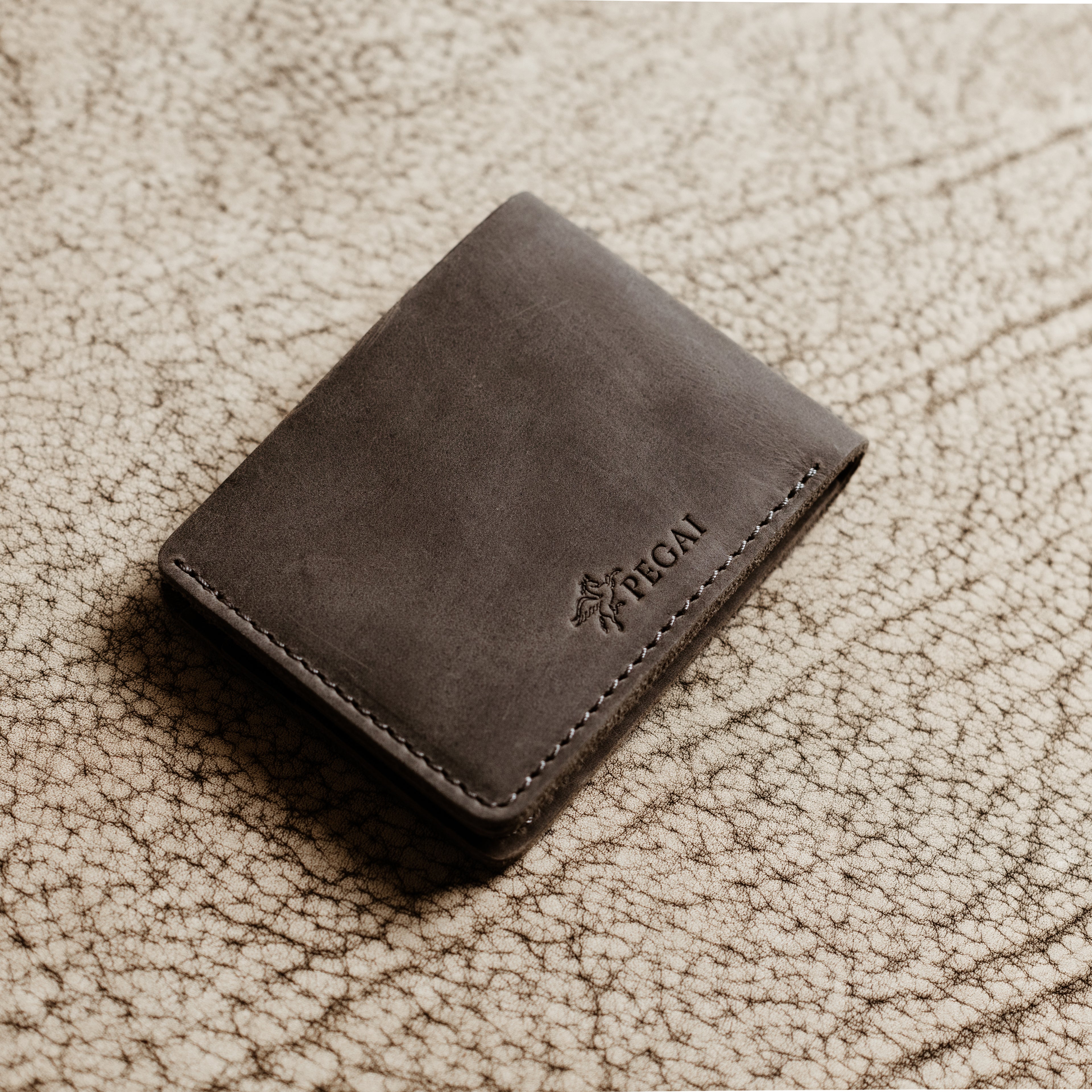 Bureau Leather Bifold Wallet in Rock Gray resting closed on a cream-toned crackled leather surface. Deep cool gray Dallas Crazy Horse calfskin exterior with natural waxy sheen and subtle tonal cloudiness. Debossed PEGAI Pegasus logo and wordmark visible on the lower right corner. Tonal gray saddle stitching along the right edge.