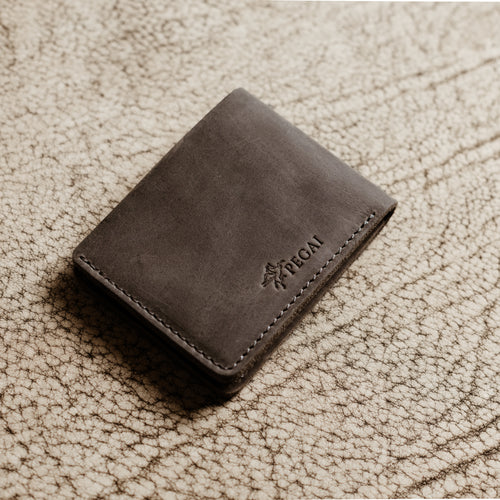 Bureau Leather Bifold Wallet in Rock Gray resting closed on a cream-toned crackled leather surface. Deep cool gray Dallas Crazy Horse calfskin exterior with natural waxy sheen and subtle tonal cloudiness. Debossed PEGAI Pegasus logo and wordmark visible on the lower right corner. Tonal gray saddle stitching along the right edge.