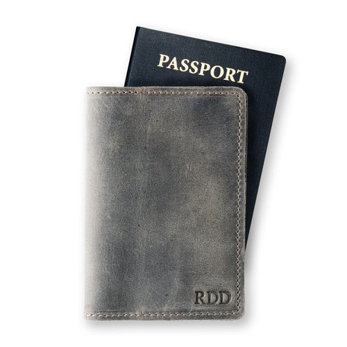 Rock gray leather passport cover in front of a navy U.S. passport on a white background. Distressed waxy surface with natural character marks. Fire-branded initials "RDD" embossed in serif font at the lower front panel. Gray hand-stitched perimeter visible along right edge.