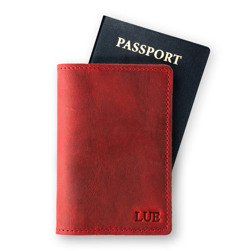 Rose red leather passport cover in front of a navy U.S. passport on a white background. Waxy crazy horse surface with natural tonal variation. Fire-branded initials "LUE" embossed in serif font at the lower front panel. Red hand-stitched perimeter visible along right and bottom edges.