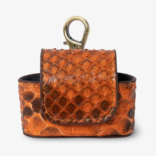 Coil AirPods case in Sunset python leather, front view. Warm amber and burnt orange scales with rich brown undertones. Magnetic flap closure and antique brass keychain clip at top. Black interior visible.